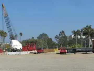 A large covered boulder for Levitated Mass sits on a flatbed trailer in a busy construction staging area with cranes, trucks, and stacked materials.