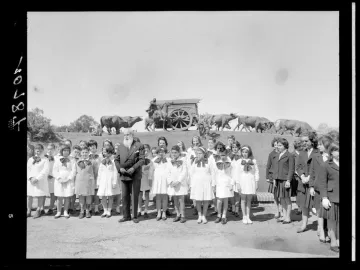 Children and an older man stand in the foreground with the Monument to the Cart on a grassy slope behind them.