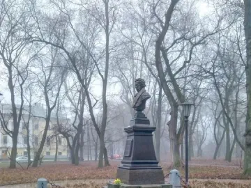 Bust of Pushkin on a tall tiered pedestal in a park with bare trees and a low chain fence.