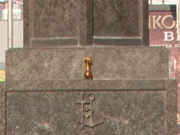 Stone base of the Statue of Sofia with a carved cross-and-anchor emblem and a small vessel on the ledge above it, partly obscured by foliage.