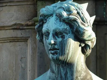 Close-up of a weathered, green-blue patinated statue head with wavy hair and a spiked headdress.