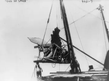 Golden Boy statue being hoisted by cables, with wooden bracing and workers below guiding the lift.
