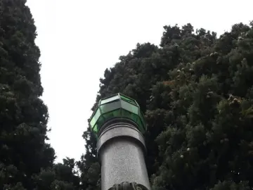 Lighthouse of Lierna, a tall stone monument with a green lantern top beside dense trees.