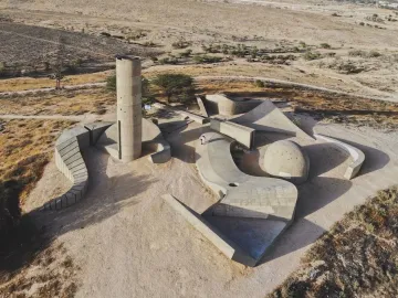 Aerial view of the Monument to the Negev Brigade, showing a concrete tower, curved walls, and geometric platforms on a dry landscape.