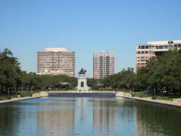 Sam Houston Statue at the end of a reflecting pool, framed by park trees and city buildings.