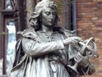 Copernicus Monument bronze statue holding a spherical instrument, with trees and a brick building behind it.