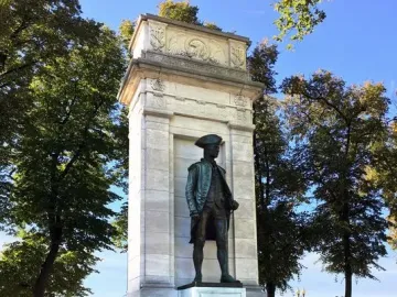 John Paul Jones Memorial with a bronze statue in a marble pylon against trees, water, and a distant bridge.