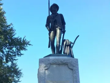 The Minute Man bronze statue stands on a stone monument with a musket, plow, and coat at the base, while a black dog sits nearby.