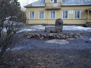 A recumbent stone lion sculpture sits on patchy ground near a yellow building, with loose rocks around it.