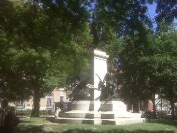 Tadeusz Kościuszko statue on a stone pedestal framed by dense green trees, with a person sitting on the steps reading.