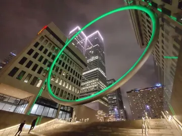The Ring glows green against illuminated skyscrapers at night, with stairs and a few blurred figures below.