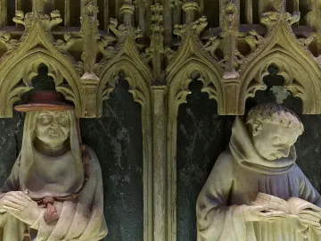 Two carved stone figures in Gothic niches from the Tomb of Charles III and Eleanor, one wearing a wide-brimmed hat and the other reading a book.