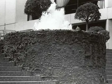 San Francisco Fountain with water cascading from its top edge in front of a modern building and two small trees.