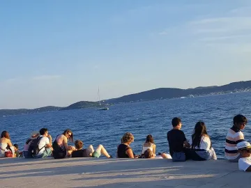 People sitting on the Sea Organ stone steps by the blue sea in Zadar, looking toward distant islands and a sailboat.