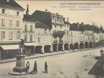 Statue of the Volunteer of 1792 on a pedestal beside a cobblestone street with arched arcades and pedestrians.