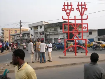 Njé Mo Yé, a large red geometric metal sculpture with two stylized heads, stands in a busy street setting.