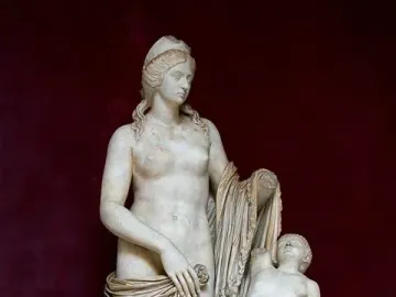 Venus Felix marble statue with a small winged child beside her, set on a inscribed plinth against a dark red background.