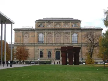 Buscando la Luz II, three tall dark reddish-brown sculptures on a green lawn in front of a classical building.
