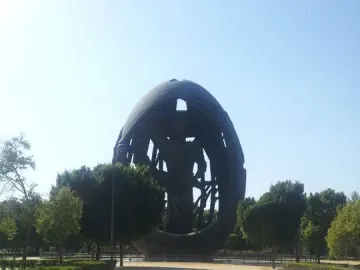 The Birth of a New Man sculpture stands at the end of a dirt path in a park, with trees and bushes around it.