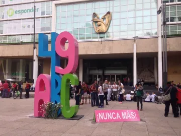 Antimonumento 49 ABC stands on a paved plaza in front of a modern building, with a small bouquet of white flowers and a pink sign reading \