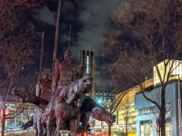 The Partisans equestrian monument at night with riders holding tall staffs, an informational plaque, and a floral wreath in front of city buildings and bare trees.