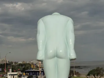 Back of the pale blue Greetingman sculpture with a cityscape, beach, and water in the background