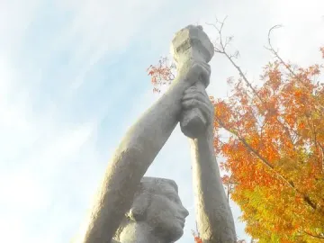 Victims of Communism Memorial statue viewed from below, with the female figure raising an object overhead against a bright sky and autumn leaves.