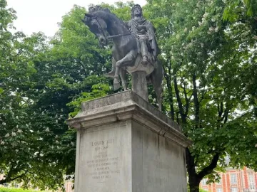 Equestrian statue of Louis XIII on a tall stone pedestal behind a black fence, with trees and a brick building in the background.