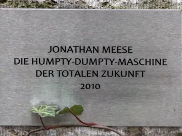 Brushed metal plaque for Jonathan Meese’s Humpty-Dumpty-Maschine mounted on a textured wall with a small ivy vine nearby.