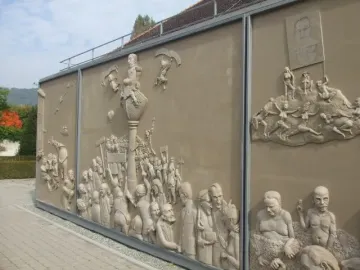Detail of Ludwigs Erbe showing a sculptural relief with a central column and crowded figures on a beige wall.
