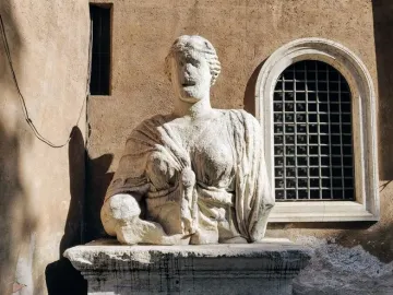 Madama Lucrezia, a weathered white stone bust of a woman on a pedestal beside a tan wall with windows.