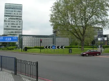 Michael Faraday Memorial on a grassy roundabout island beside roads and traffic lights, with buildings and trees in the background.