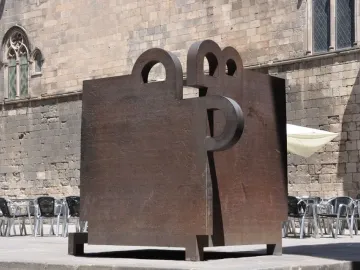 Topos V by Eduardo Chillida in front of a stone building with a pointed arch window and café tables nearby.
