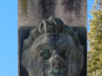 Weathered bronze bust of Julius Fučík set into a rough stone stele, with verdigris against a blue sky.