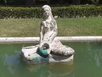Weathered stone mermaid statue in a rectangular pool with murky green water, holding a turquoise object in her lap.