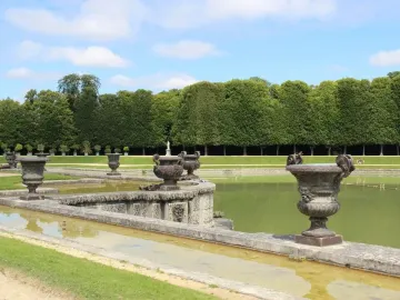 Neptune Fountain at Versailles with a large basin, decorative stone urns, and a trimmed tree hedge under a blue sky.