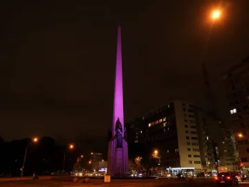 The Obelisk of Montevideo lit in purple at night, with buildings and traffic around it.