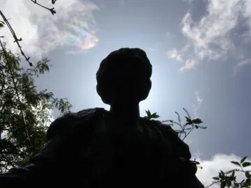 Dark silhouetted bust of the Pankhurst Memorial against a bright sky with wispy clouds and tree branches.