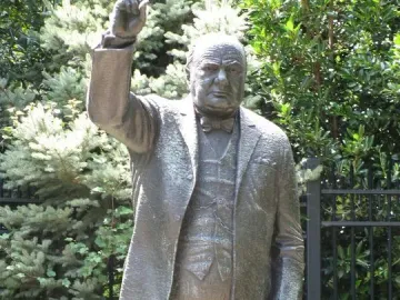 Bronze Winston Churchill statue standing outdoors, raising his right hand in a V-sign and holding a walking stick.