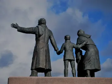Auswandererdenkmal sculpture of three dark metallic figures standing together on a stone pedestal against a blue sky.