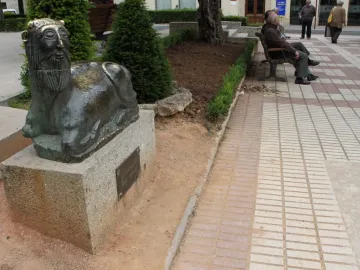 Bicha of Balazote sculpture on a stone plinth in a park, with a reclining animal body and human-like bearded head.