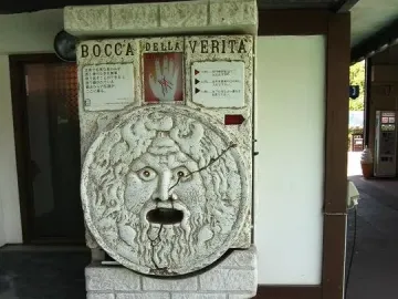 Bocca della Verità stone face replica with wide-open eyes and a gaping mouth, shown on a textured pedestal with a Japanese sign above it.