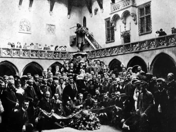 Copernicus Monument on a pedestal beside a large floral wreath, surrounded by a crowd in a grand hall.