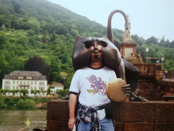 A man poses inside the face opening of the Heidelberg Bridge Monkey sculpture, with a bronze monkey holding a disc on a brick wall behind him.