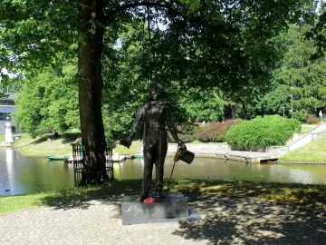 Bronze Pushkin statue on a stone plinth beside a canal, with flowers at the base and a large tree overhead.
