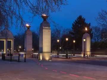 Commonwealth Memorial Gates at dusk, with a domed pavilion, four lit stone pillars topped by flames, and bare trees against a blue sky.