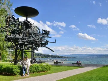 Heureka sculpture standing on a grassy lawn beside Lake Zurich, with mountains in the distance.