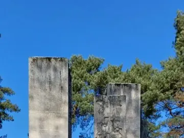Tall stone steles of the Julius Fučík Memorial stand in a snowy park with evergreen trees and nearby buildings.