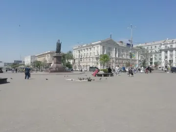 The Monument to Dimitry of Rostov stands on a tiered pedestal in a city square, with pedestrians, pigeons, and buildings in the background.
