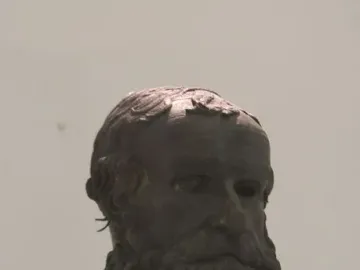 Bronze head of the Porticello Philosopher with a curly beard and worn surface on a dark stand.